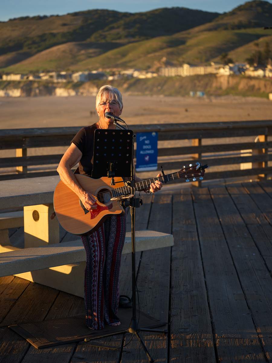 Joelyn Lutz performing live on the Pismo Beach pier at sunset, singing into a microphone with her acoustic guitar