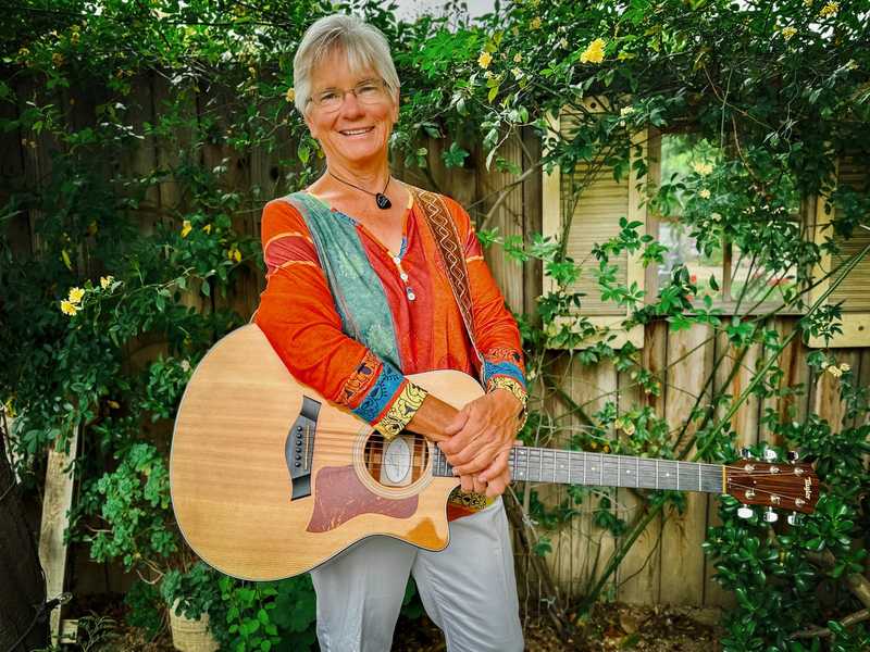Joelyn Lutz standing in a garden holding her Taylor acoustic guitar, smiling, in an orange and teal patchwork top