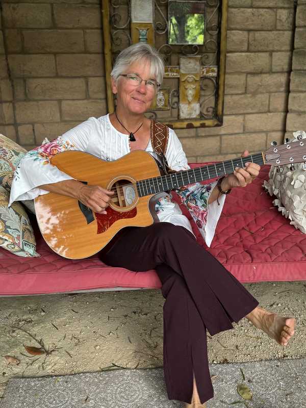 Joelyn Lutz press photo, 2023: playing guitar barefoot on a red daybed against a brick wall