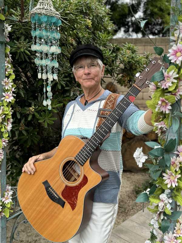 Joelyn Lutz press photo, 2023: standing under a flowered garden arch with her guitar, wearing a striped blue sweater and black cap