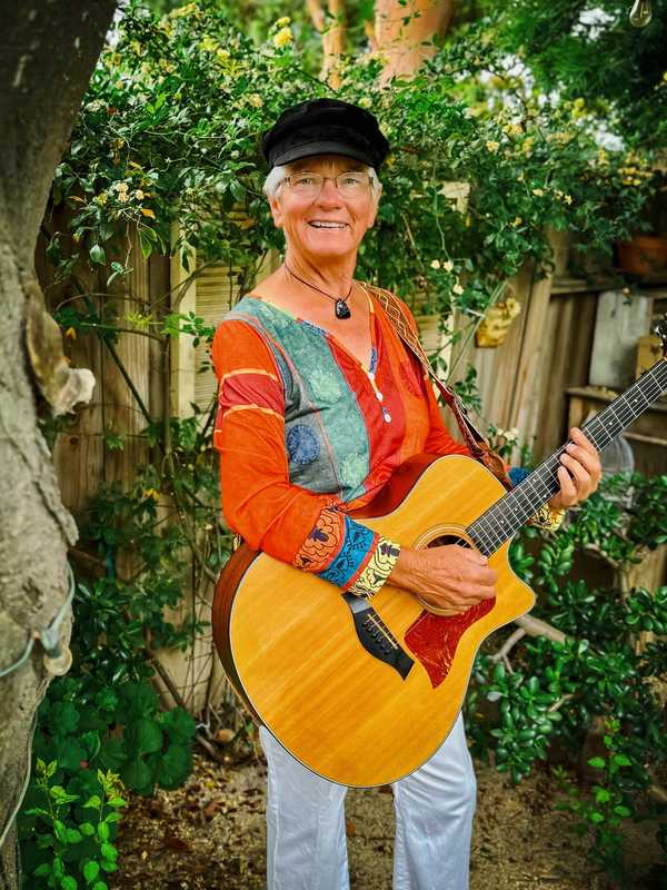 Joelyn Lutz press photo, 2023: laughing in a black cap with her acoustic guitar in front of climbing yellow roses