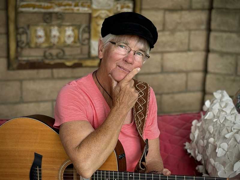 Joelyn Lutz press photo, 2023: seated on a patio daybed in a pink shirt and black cap, chin resting on her hand, guitar across her lap